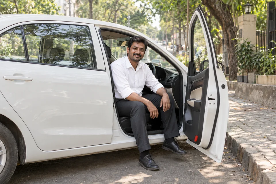 A Captain seated comfortably in the open doorway of his parked white cab on a leafy city street.